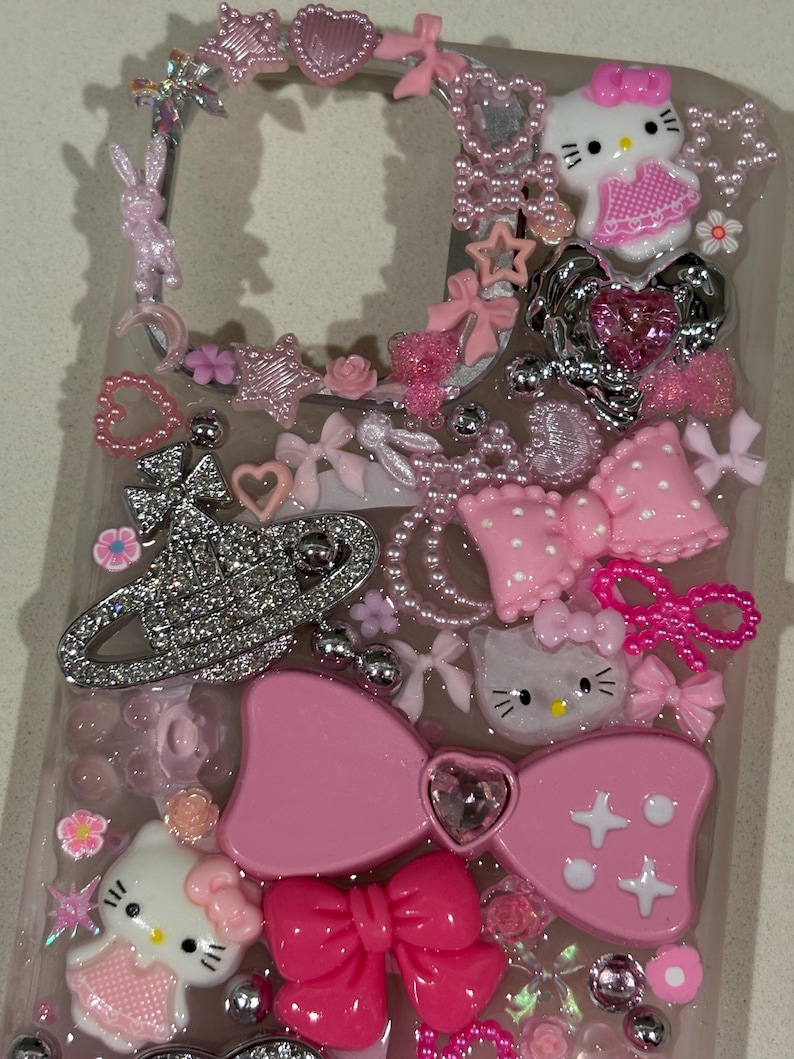 K&ouml;nnte beinhalten: Eine transparente Handyh&uuml;lle, verziert mit einer Vielzahl von rosa Verzierungen. Enth&auml;lt Hello Kitty-Figuren, Schleifen, Herzen, Sterne und ein glitzerndes silbernes Emblem. Die H&uuml;lle ist mit dekorativen Elementen in verschiedenen Texturen und Gr&ouml;&szlig;en verziert.