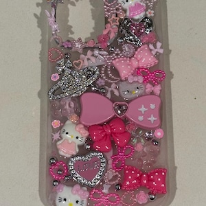 K&ouml;nnte beinhalten: Transparente Handyh&uuml;lle mit rosa Schleifen, Herzen und Hello Kitty-Figuren. Die H&uuml;lle ist mit Strasssteinen und silbernen Perlen verziert und schafft eine verspielte und m&auml;dchenhafte &Auml;sthetik. Das Design integriert mehrere Rosat&ouml;ne.