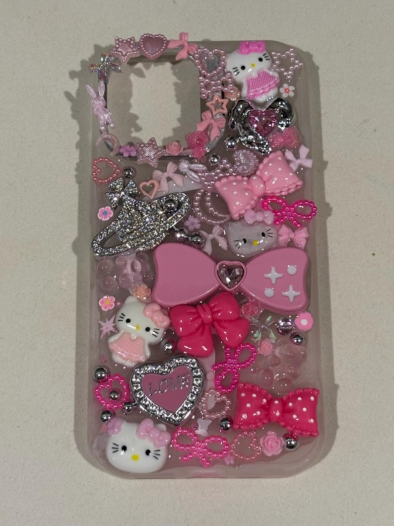 K&ouml;nnte beinhalten: Transparente Handyh&uuml;lle mit rosa Schleifen, Herzen und Hello Kitty-Figuren. Die H&uuml;lle ist mit Strasssteinen und silbernen Perlen verziert und schafft eine verspielte und m&auml;dchenhafte &Auml;sthetik. Das Design integriert mehrere Rosat&ouml;ne.