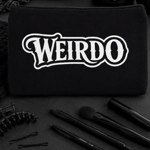 May include: A black rectangular zippered pouch featuring the word WEIRDO printed in large white stylized letters across the center. The pouch is accompanied by a black hair claw clip, two black bobby pins, two makeup brushes, and a black tube.