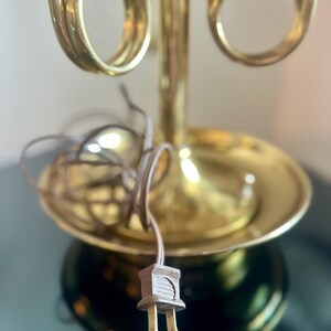 May include: A vintage brass table lamp with a decorative design. The lamp features a round base, a flared shade holder, and a curved, horn-like detail. The electrical cord and plug are visible.