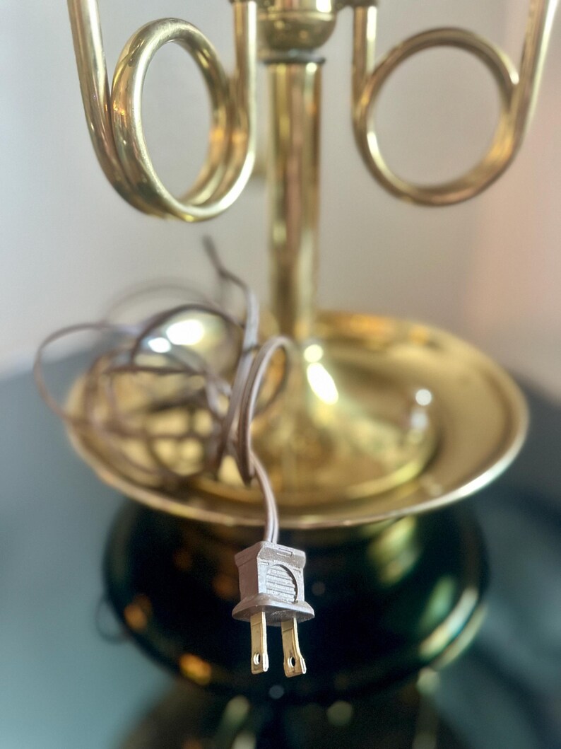 May include: A vintage brass table lamp with a decorative design. The lamp features a round base, a flared shade holder, and a curved, horn-like detail. The electrical cord and plug are visible.