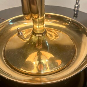 May include: Close-up of a brass lamp base with a circular dish-like element. The lamp has a polished, reflective surface, with a switch on the side. The base is set on a dark, glossy surface.