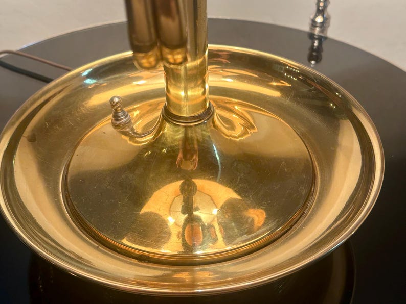 May include: Close-up of a brass lamp base with a circular dish-like element. The lamp has a polished, reflective surface, with a switch on the side. The base is set on a dark, glossy surface.