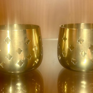 Puede incluir: Dos candelabros votivos de metal dorado. Cada uno tiene una forma redondeada con una serie de recortes en forma de trébol. Los candelabros reflejan la luz, creando un brillo cálido. Estas piezas decorativas son ideales.