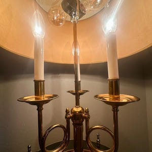 May include: A brass candelabra-style lamp with three light bulbs and three candle-shaped lights. The lamp has a gold-colored base and a beige lampshade. The bulbs are lit, casting a warm glow.