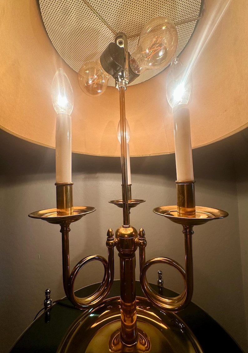 May include: A brass candelabra-style lamp with three light bulbs and three candle-shaped lights. The lamp has a gold-colored base and a beige lampshade. The bulbs are lit, casting a warm glow.