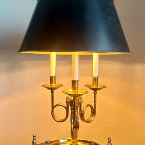 May include: A brass candelabra-style table lamp with a black lampshade. The lamp features three candle-shaped light bulbs and a decorative brass base. The lamp is on a black table.