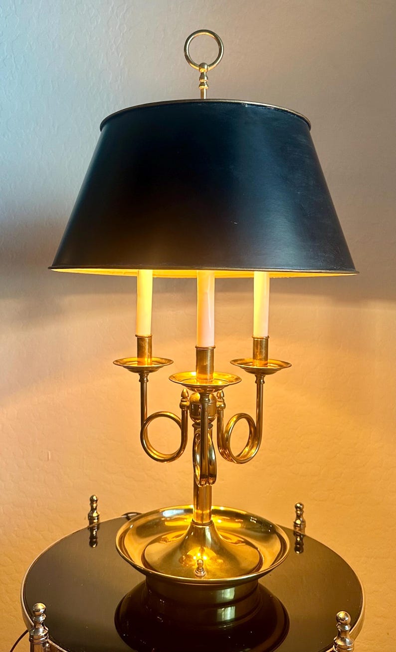 May include: A brass candelabra-style table lamp with a black lampshade. The lamp features three candle-shaped light bulbs and a decorative brass base. The lamp is on a black table.