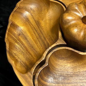 Puede incluir: Una bandeja de madera para servir con múltiples compartimentos. La bandeja tiene un acabado natural de vetas de madera y una tapa en forma de calabaza. Los compartimentos tienen forma de hojas y corazones. La bandeja está diseñada para servir aperitivos.