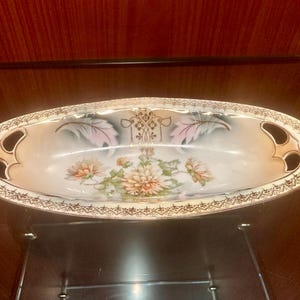 May include: Oval-shaped porcelain dish with a floral design. The dish features a cream-colored interior with a painted floral arrangement of peach and green flowers. The rim is trimmed with gold detailing, and it has two handles. The dish is displayed on a glass shelf.