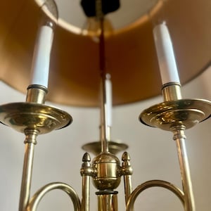 May include: A brass candelabra with three arms, each holding a white candle. The candelabra has a gold lampshade and a decorative design. The overall aesthetic is vintage and elegant, suitable for home decor.