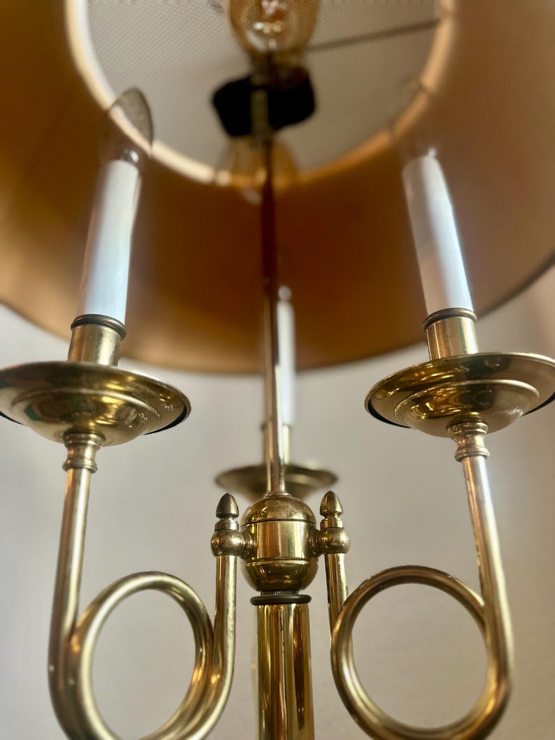 May include: A brass candelabra with three arms, each holding a white candle. The candelabra has a gold lampshade and a decorative design. The overall aesthetic is vintage and elegant, suitable for home decor.
