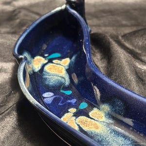 Puede incluir: Una taza de cerámica blanca con asa negra y el texto "But First, Coffee" en negrita negra. La taza se encuentra sobre un fondo borroso, sugiriendo un ambiente acogedor.