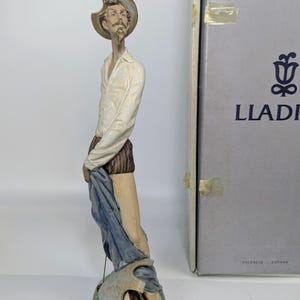 May include: A tall, slender porcelain figurine of a man with a goatee, wearing a wide-brimmed hat, a white collared shirt, and dark shorts. He stands beside a stack of books, draped in a blue cloth, with a Lladró box visible in the background.