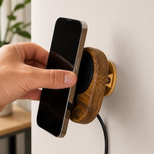 Könnte beinhalten: Ein wandmontiertes kabelloses Telefonladegerät mit einem ovalen Gehäuse aus dunklem Holz, einem schwarzen kreisförmigen Zentrum und einer goldfarbenen runden Basisplatte. Ein schwarzes Stromkabel führt aus der Holzeinheit an der Wand.