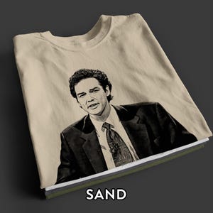 May include: A sand-colored t-shirt featuring a black and white graphic illustration of a person with dark hair wearing a suit, white collared shirt, and a patterned tie. The word SAND is printed in white capital letters at the bottom of the image.