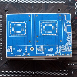 May include: A rectangular electronic component featuring a vibrant blue printed circuit board surface with two distinct square contact pad arrays and intricate white circuitry patterns, mounted on a dark brown motherboard base.