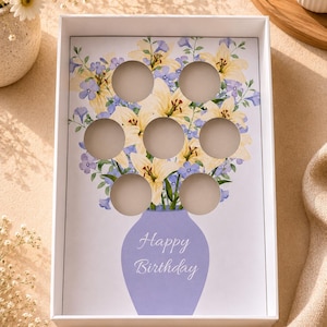 May include: White gift box with a floral design and the words "Happy Birthday." The interior features a floral arrangement of yellow lilies and blue flowers, with seven circular cutouts. The vase is a light purple colour.