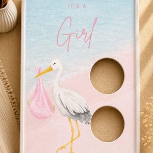 Peut inclure: Un cadre blanc rectangulaire contenant une impression avec une cigogne aquarelle portant un baluchon rose. Le texte IT'S A Girl est imprimé en rose. Le fond présente un dégradé bleu et rose doux. Deux découpes circulaires sont sur la droite.