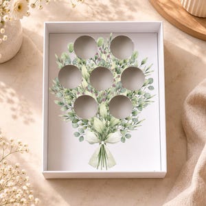 May include: A white gift box with a floral bouquet design. The box interior features seven circular cutouts. The bouquet illustration includes green leaves, purple accents, and a light green ribbon. The box is on a light-colored surface.