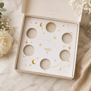 May include: An open, off-white box with a decorative interior featuring the word "ARIES" and a zodiac symbol. The box has circular cutouts and gold star and crescent moon accents. The box is on a marble surface.