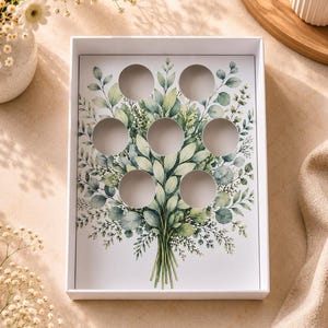 Puede incluir: Una caja de regalo blanca con un diseño botánico de hojas verdes y siete recortes circulares. La caja está vacía, con un fondo neutro y una iluminación suave.