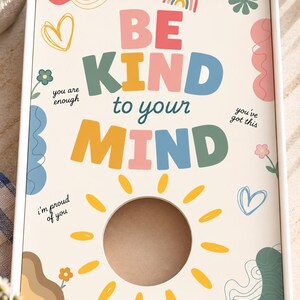 May include: A rectangular white gift box with a cream-colored lid featuring the text BE KIND to your MIND in colorful block letters. The design includes a yellow sun graphic with a circular cutout, small flowers, hearts, and the phrases you are enough, you've got this, and i'm proud of you.