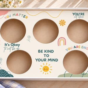 May include: A rectangular white cardboard box with five circular cutouts, featuring colorful illustrations of flowers, rainbows, and a sun, along with the text: YOU MATTER, It's Okay to not be ok, YOU'RE not alone, BE KIND TO YOUR MIND, and YOU ARE ENOUGH.
