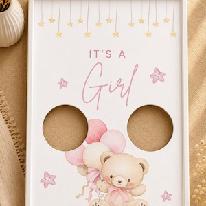 Peut inclure: Un cadre rectangulaire blanc contenant une impression avec le texte IT'S A Girl en rose, illustrant un ours en peluche marron clair tenant des ballons roses et blancs, entouré de petites étoiles roses et d'étoiles jaunes suspendues, avec deux découpes circulaires.