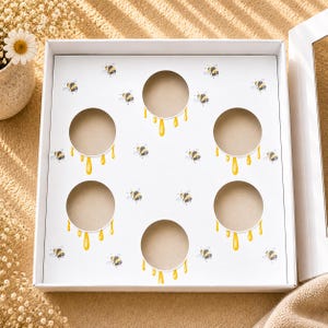 Puede incluir: Caja de cartón blanca con seis recortes circulares. La caja está decorada con abejas de acuarela y gotas de miel amarillas. La caja está abierta, revelando el interior. El tema es la miel y las abejas.