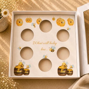 Puede incluir: Una caja blanca con interior crema con recortes circulares. El interior está decorado con ilustraciones de acuarela de panales, abejas y barriles de miel. También está presente el texto "What will baby bee...".