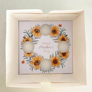 May include: A square gift box with a floral design. The box contains a card with a sunflower wreath and the text "Happy Mother's Day". Five round, white objects are arranged on the card.