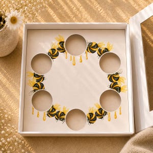 Puede incluir: Una caja blanca con un interior decorativo que presenta un anillo de abejas de acuarela con alas amarillas y rayas negras y amarillas. La caja tiene recortes circulares y un diseño de goteo de miel. Un pequeño jarrón con flores blancas está en el fondo.