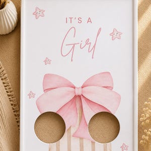 Peut inclure: Un cadre rectangulaire blanc contenant une impression avec le texte IT'S A Girl en lettres roses. Le design présente un grand nœud rose de style aquarelle, des rayures beiges verticales et plusieurs petites étoiles roses souriantes.