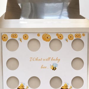 Puede incluir: Portador de cupcakes de cartón blanco con 12 recortes circulares. El borde superior presenta ilustraciones de panales amarillos y naranjas. La frase "What will baby bee..." está escrita en escritura dorada, con un dibujo de abeja.