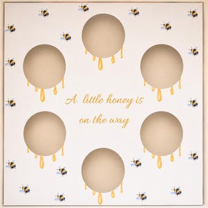 Puede incluir: Caja blanca con cinco círculos beige, cada uno con una gota de miel amarilla. La caja está decorada con abejas de acuarela. El texto "A little honey is on the way" está escrito en oro.