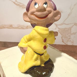 以下が含まれることがあります： 白雪姫のキャラクター、ドーピーの陶器製フィギュア。光沢のある黄色いチュニックに2つの赤いボタン、紫色の帽子、大きな耳、茶色の靴が特徴です。明るい肌色で、大きく口を開けて微笑み、大きな青い目が上を向いています。
