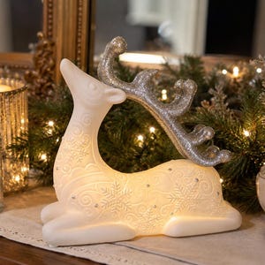 May include: A white, illuminated deer figurine featuring embossed snowflake patterns and small silver accents across its body. The deer is in a reclining position and has large, textured silver glitter antlers that curve backward over its back.