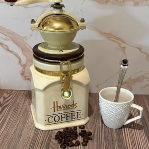 Op de afbeelding: Een crèmekleurige keramische koffiebus met een goudkleurige metalen sluiting en een handmatige molen bovenop, voorzien van het Harrods Knightsbridge Coffee logo. Daarnaast staat een witte, textuurrijke keramische beker met een zilveren lepel.