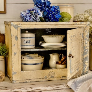 May include: A rectangular metal pie safe with a distressed cream and gray paint finish, featuring a hinged door with a metal latch. Inside are two shelves holding a white ceramic cake jar, a stack of white bowls, a pitcher, and a ceramic bowl.