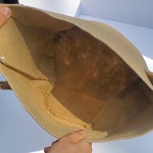 Può includere: Una borsa tote beige di forma rettangolare con un'ampia apertura. La borsa è realizzata in un materiale ruvido e strutturato e ha un design semplice. L'interno della borsa è visibile, mostrando lo stesso materiale beige.