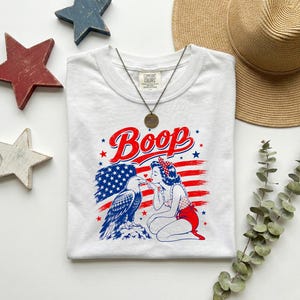 May include: A white t-shirt featuring a graphic print of a person with a red polka dot headband kneeling next to a bald eagle. The design includes the text Boop and a red, white, and blue American flag background. A metal pendant necklace is included.