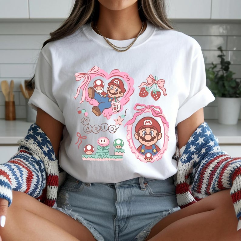 Op de afbeelding: Wit T-shirt met een kleurrijke afbeelding van Mario uit Super Mario Bros. Het ontwerp bevat roze harten, paddenstoelen, aardbeien en het woord "MARIO". Het shirt heeft korte mouwen en een ronde hals.