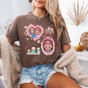 Op de afbeelding: Een bruin t-shirt met een kleurrijk grafisch ontwerp van Mario-personages in roze frames en hartvormen. Het ontwerp bevat aardbeien, paddenstoelen en de letter "M". Het shirt wordt gedragen door een persoon die een blauwe spijkerbroek draagt.