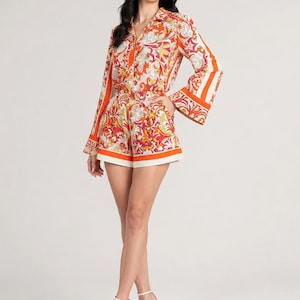 May include: A two-piece set consisting of a long-sleeved button-down shirt and matching shorts. The garment features a vibrant orange, red, yellow, and white paisley-inspired pattern with solid orange and white border accents on the cuffs and hem.