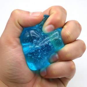 Puede incluir: Una mano apretando una sustancia clara y azul gelatinosa. El gel tiene pequeñas burbujas y es translúcido. El fondo es blanco, lo que enfatiza la textura y el color del gel.