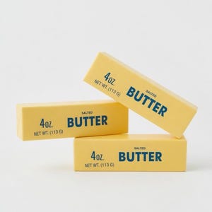 Op de afbeelding: Drie gele boterpakjes, elk met de tekst "4 oz. NET WT. (113 g)" en "SALTED BUTTER" in blauwe letters. De boterpakjes zijn gestapeld tegen een witte achtergrond.