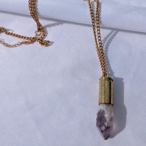 May include: A gold-colored chain necklace with a pendant. The pendant features a raw, purple amethyst crystal encased in a gold-toned metal casing. The necklace is displayed on a white background.