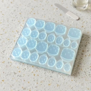 May include: A sheet of pale blue toe nail adhesive pads. The pads are arranged in rows and columns, with varying sizes to fit different toes. A nail file and a bottle of clear liquid are in the background.